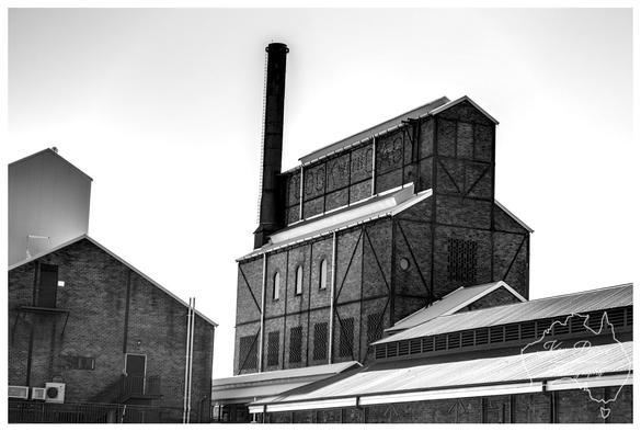A high contrast black and white photograph of the historic Launceston Gasworks buildings. The central structure is a large, dark brick industrial building with external steel framing and a tall, slender chimney rising from the top. 

Smaller brick and corrugated iron buildings are visible on either side and in the foreground. The bright white sky provides a dramatic background.