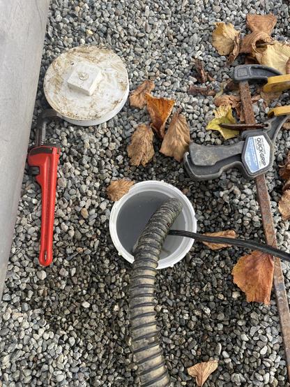 looking down into some gravel with some fallen brown leaves in the middle of the picture is a open 4 inch pipe with water nearly at the top of it and a smaller corrugated pipe and electrical cable going down in. There is a red handled pipe wrench beside and the cap is just above it.