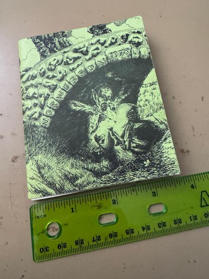 A green booklet next to a green plastic ruler showing that the booklet is about 3.5 inches wide by 4 inches tall. It has a cover with artwork of an elfin creature sitting by a stream under a bridge with 'Norwescon 1978' inscribed on the stones, smoking a pipe and writing on a parchment with a large feather quill.