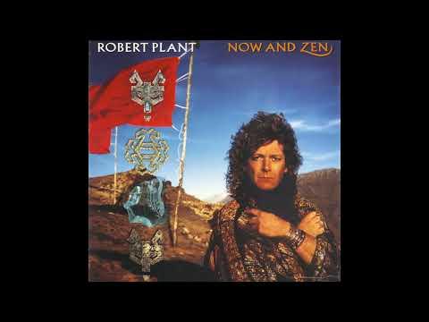 An album cover showing a middle-aged white man with bronze-colored curly hair (Robert Plant of Led Zeppelin) wearing a cloak with bronze/gold embellishments, with his arms crossed. He is standing in a mountainous desert under a blue sky with a few wispy clouds. Next to him is a flagpole with a red flag (with a wolf-like symbol), with other symbols underneath.
Text: ROBERT PLANT      NOW AND ZEN