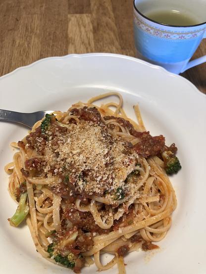 A plate of pasta topped with a meat and vegetable sauce, garnished with breadcrumbs. A cup of green tea is visible in the background.