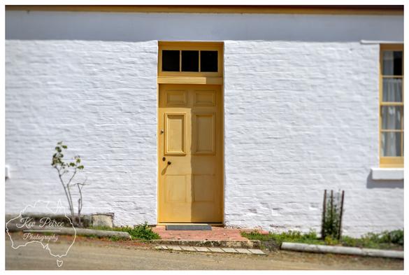 A historic white painted brick wall with a single, brightly painted mustard yellow wooden door in the center.

The door has a rectangular window transom above it, also painted yellow. A small step leads up to the door from a dirt path in the foreground.