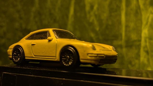 Zu sehen ist ein gelber Porsche 911 Carrera (Siku) vor grünlich-gelblichem Hintergrunz