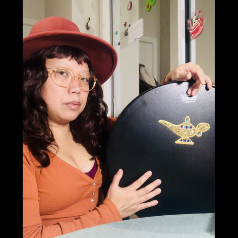 Me wearing orange hat, glasses, top holding a 16” black Remo shamanic/ Buffalo drum with magic lamp sticker
