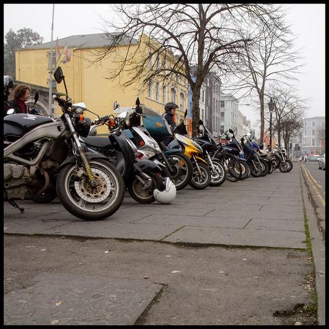 Row of motorcycles parked along wide pedestrian pavement on Grand Parade Cork with riders standing beside bikes, yellow and grey period buildings visible behind bare winter trees on overcast day, photo from 20 years ago showing former motorcycle parking arrangement