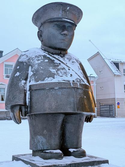 Das Foto zeigt die Statur "Toripolliisi" in Oulu in Finnland.
Es ist die Figur eines recht beleibten Polizisten.
Im Hintergrund sind ein paar Häuser zu sehen.
Der Boden ist schneebedeckt, auf der Statur liegen ebenfalls Schneereste.