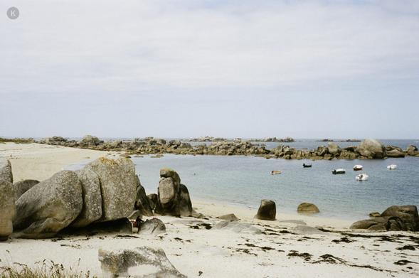 Ruhige Küstenbucht mit hellem Sandstrand, großen Granitfelsen im Vordergrund und mehreren kleinen Booten auf dem klaren, blauen Wasser unter leicht bewölktem Himmel.