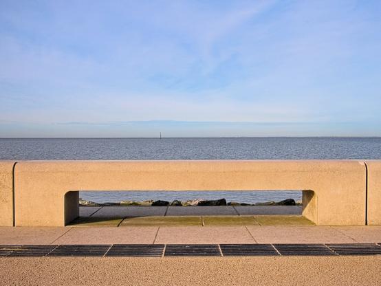 We are at the seaside. In the foreground is a flat concrete bench. It is illuminated by the sunlight, which gives the concrete a warmer appearance. The sea can be seen beneath the bench and, of course, behind it. It is high tide.