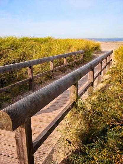 Ahead of us lies a path with a wooden railing. To the left and right, grass grows on low dunes. At the end of the boardwalk, the sand begins, and beyond it lies the sea. It is a sunny day; the sky and sea are blue.