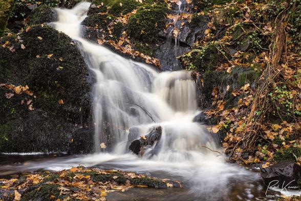 Photographie en couleur présentant une chute d'eau d'environ 1,50 m sur un petit ruisseau entre de gros rochers couverts de mousse et des nombreuses feuilles tombées des arbres voisins. 
La durée d'une seconde de l'exposition a généré un filé de l'eau dégringolant des rochers dans un petit bassin avant de continuer, hors cadre, ça descend du coteau via une succession de ressauts dont vous pourrez voir quelques-uns sur la seconde photo.