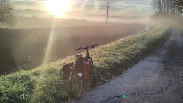 Ein schwarzes e-bike auf einem Asphaltweg, links ein grasbewachsener Deich, tief links darunter die Weschnitz. Rechts im Hintergrund Bäume, links Felder und dahinter in Nebelschwaden die Bergstraße. Dahinter die aufgehende Sonne.