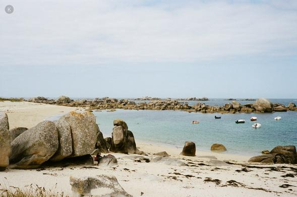 Ruhige Küstenbucht mit hellem Sandstrand, großen Granitfelsen im Vordergrund und mehreren kleinen Booten auf dem klaren, blauen Wasser unter leicht bewölktem Himmel.