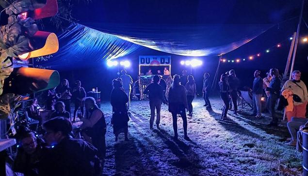 Konzertevent im Freien bei Nacht. Menschen tanzen vor einer Bühne mit DJ-Pult und Lichttechnik. Dunkler Himmel, blaues Zeltdach, gedämpftes Licht.