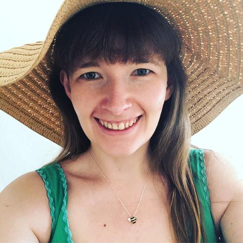 A headshot of a white woman with reddish-brown hair and a fringe, smiling straight forwards at the camera and wearing a straw sun hat and a green strappy dress.