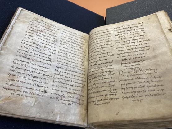 An open ancient manuscript featuring handwritten text on aged parchment pages. The script appears to be in Latin, with several lines neatly organized in columns. The book is displayed on a dark surface.