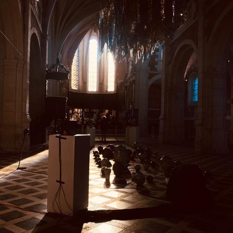The photograph shows the interior of a large church. Sunlight enters through tall stained-glass windows on the left, illuminating the checkered stone floor. A reflective installation made of metallic or golden material hangs horizontally from the ceiling near the windows. On the right side of the image, several rounded objects—possibly vases or glass vessels—are placed on the floor, reflecting the light. In the foreground, there is a white rectangular pedestal with black tape and electrical cables attached to it. The architecture includes high arches, ribbed vaults, and stone columns. The lighting creates long shadows across the floor.
