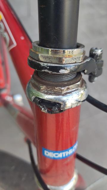 Gros plan sur la direction du vélo rouge.
Les écrous de la partie haute du jeu de direction sont en partie démontés et piqués de rouille, la cuvette supérieure est cassée laissant apparaître la cage des billes déformée