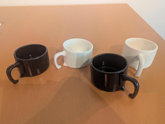 Vier Tassen, zwei schwarz und zwei weiß, stehen auf einem Wohnzimmertisch in hellem Holz. Die Tassen wirken, als würden sie in der Tischplatte versinken.