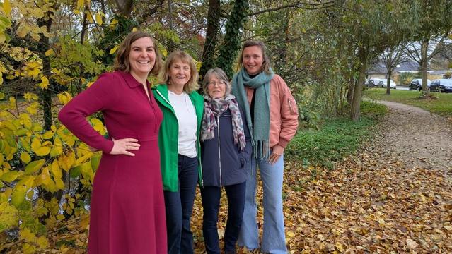 Drei Frauen stehen lächelnd auf einem kiesbedeckten Weg im Herbstlaub. Eine trägt ein rotes Kleid, eine grüne Jacke und die dritte eine blaue Jacke mit Schal.