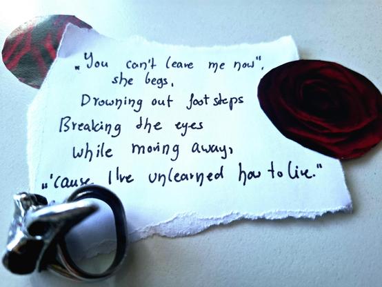 A piece of paper with unevenly torn edges on a white surface. Red paper roses on both sides, and a raven skull ring. 

The handwritten lines are:

“You can’t leave me now”,
She begs,
Drowning out footsteps
Breaking the eyes
While moving away,
“’cause I’ve unlearned how to live.”