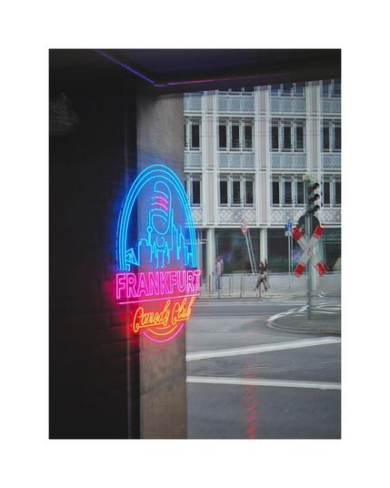 A vertical photo taken from outside, looking through a window pane. The main subject is a brightly lit neon sign for the "Frankfurt Comedy Club," which is visible inside the establishment (on the left, against a dark brick wall). The sign glows in blue and pink/red, depicting the Frankfurt skyline with a microphone. The window's glass creates a significant reflection of the surrounding streetscape, including a railroad crossing sign (St. Andrew's cross) and a tall, multi-story building with a patterned facade, blending the exterior and interior views.