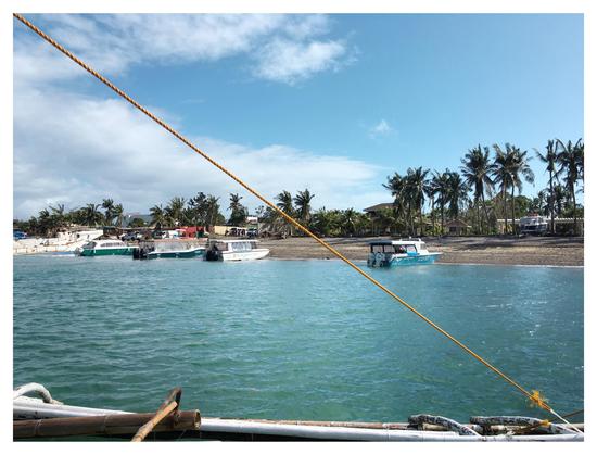 Photo of an island beach as seen from an approaching boat: coconut trees, blue sky with white clouds, boats anchored near the shore. A taut yellow rope, fastened to a boat outrigger at bottom frame, stretches from the upper left corner to the lower right corner of the photo and diagonally bisects the view.
