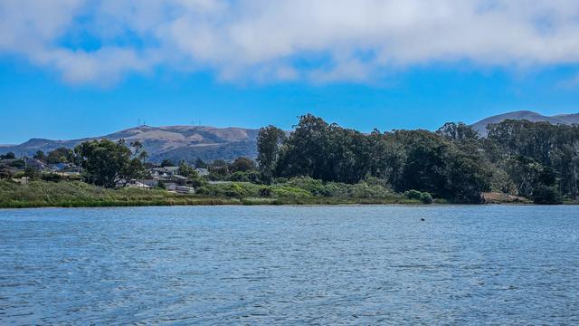 A landscape photograph depicts a body of water in the foreground with a shoreline of vegetation and a mountainous backdrop under a clear blue sky. The water is a shimmering blue with subtle ripples, extending across the majority of the frame. A dense line of green trees and shrubs hugs the shoreline, with a few houses visible through the foliage on the left side. In the distance, rolling brown mountains rise, partially obscured by a light haze, with a single, tall antenna or tower visible on the peak of the furthest mountain. The sky is a vivid blue with scattered white clouds, suggesting a sunny day.