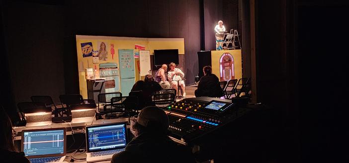 Photo of stage set up for the play called Trash. Photo taken behind the production station with black box theater style set up. Two Deaf actors are seated on the stage surrounded by bright set while one actor is standing on the left on a podium representing the jukebox dressed in a shiny silver outfit with overhead spotlight on them. The rest of the stage is covered in dim lights with spotlights focusing on the actors.