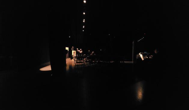 Panormaic photo of stage set up for the play called Trash. Photo taken from side of the stage with black box theater style set up. Two  actors are standing n the stage surrounded by bright set while one actor is standing on the left on a podium representing the jukebox dressed in a shiny silver outfit with overhead spotlight on them. The rest of the stage is covered in darkness with spotlights focusing on the actors