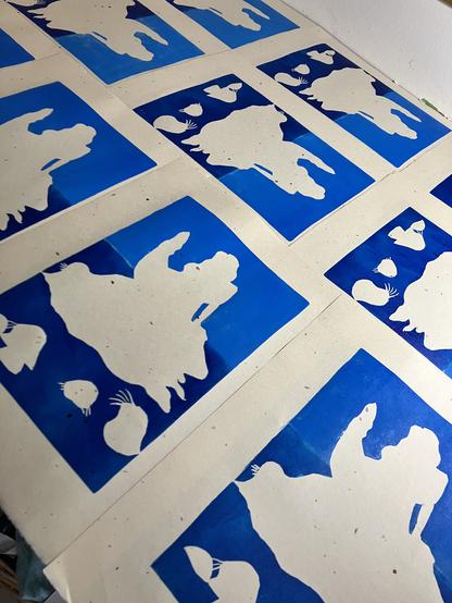 Ten of my 11”x14” linocut prints on cream coloured washi paper with bark inclusions are laid out on a table to dry. They are in their first state, with the sea and sky background printed. The outline of sea faces is unprinted in the dark blue sea below the as-of-yet-unprinted Selkie on a rock and the blue sky.
