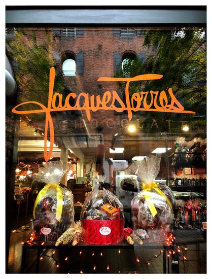 Photo of the window of a chocolate shop, featuring a table display of three baskets of chocolates in cellophane bags tied shut with ribbons and ringed with fairy lights. Above is the store name, “Jacques Torres Chocolate,” in stylized orange font. Beyond is a glimpse of the store interior with counters and more chocolate displays. The windowpane also reflects a blurry image of the scene outside: green trees in front of a red brick building with arched windows flanked by open black shutters.