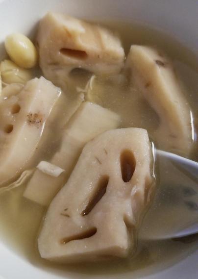 Close-up shot of a white bowl filled with a soup-like broth, showcasing large pieces of light-brown lotus root, and light-yellow ginkgo nuts. The lotus root slices feature multiple holes and a light brown hue, indicating they've been cooked, with a few visible specks on some pieces. The broth appears clear and slightly opaque, and a white spoon rests partially within the bowl.