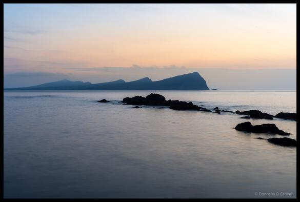 Twilight seascape showing silhouetted Three Sisters rock formations rising from calm sea with dark rocky outcrops in foreground and pastel pink and blue sunset sky above, viewed from Dingle Peninsula, County Kerry.