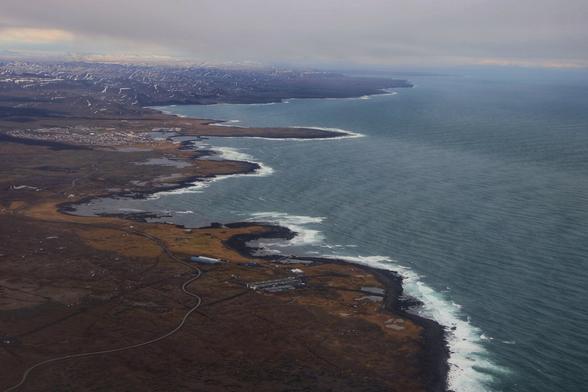 A breathtaking aerial view of a rugged coastline, where expansive, barren landscapes meet the deep blue ocean. The coastline is characterised by jagged cliffs and rocky outcrops, with patches of green and brown terrain stretching inland. Small clusters of buildings and roads are scattered across the landscape, indicating sparse human habitation. The coastline curves gently, leading the eye towards a distant horizon where the land gradually fades into the sea. In the background, a faint outline of snow-capped mountains is visible, adding depth and contrast to the scene. The sky above is overcast, casting a soft, muted light over the entire landscape and enhancing the serene and remote atmosphere.