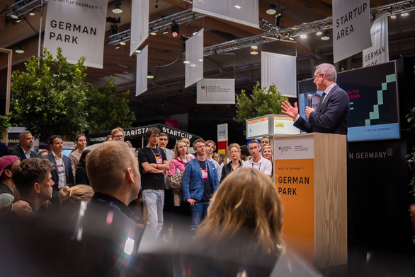 Karsten Wildberger steht vor zuhörenden Menschen an eine Redepult und spricht in ein Handmikrofon.