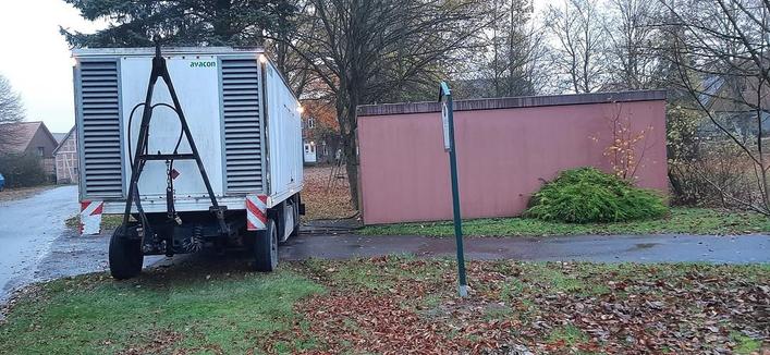 Grauer Transporter mit Leiter auf Anhänger steht auf einem Grünstreifen vor einer roten Mauer.