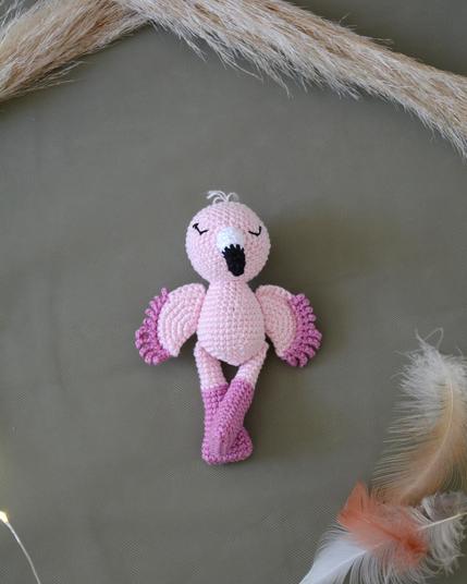 Flamant rose crocheté aux tons rose pâle et fuchsia, allongé sur un fond vert kaki. Autour, quelques plumes et tiges de pampa apportent une atmosphère douce et légère.