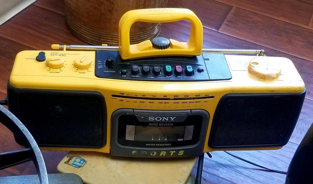 A close-up of a bright yellow Sony sports boombox, model CFS-930, with a built-in cassette player and radio. The boombox features black speakers on either side, labeled "SONY AUTO REVERSE WATER RESISTANT SPORTS" and a dial that reads "FM/AM STEREO CASSETTE-CORDER CFS-930", with multiple buttons and controls including "PHONES", "VOLUME", "TONE", "FUNCTION", "PAUSE", "PLAY", and "REC STOP/EJECT". An extendable antenna and a bright yellow handle are also present.