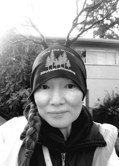 A black and white selfie of a woman wearing a black beanie with faux deer antlers and the text "Save Old Growth" above an image of three pine trees, and "#WorthMoreStanding" below. The woman has a long braid and is wearing a black jacket and a white jacket. A house and foliage are in the background.