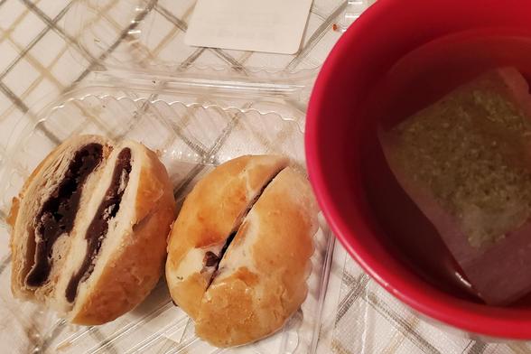 Two halved, golden-brown baked buns with red bean filling sit in a clear plastic container, beside a red mug holding a tea bag, on a beige plaid tablecloth. The buns are cut open to reveal layers of dough and dark red filling. The tea bag is visible in the mug with water.