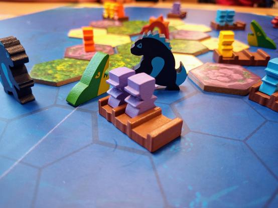 Das blaue Spielbrett mit den schönen Spielmaterialien. Braune Flöße mit Platz für drei Meeples, ein grüner Hai, der aus dem Wasser (aka Spielbrett) herausschaut, ein Gozillaartiges Monster sowie die Inselplättchen.