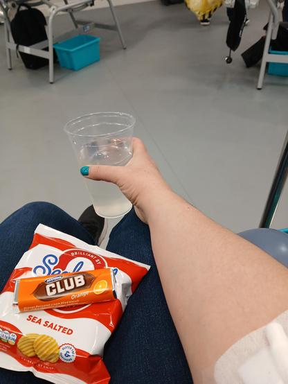 Photo of my arm and lap, with a plaster on the inside of my elbow, holding a plastic cup of squash and on my lap is a packet of Ready Salted Seabrook's and an Orange Club chocolate biscuit