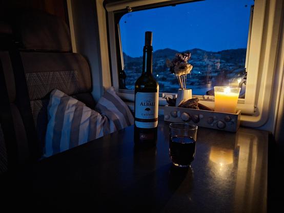 Ein Stillleben, das in einer dunklen Umgebung aufgenommen wurde, Innenraum eines Campervans, mit einer hellen Aussicht durch ein rechteckiges Fenster. Im Vordergrund befindet sich eine reflektierende dunkle Oberfläche, auf der ein Glas mit rotem Wein und eine Flasche Wein stehen. Ein Glashalter mit einer einzelnen Kerze befindet sich in der oberen rechten Ecke. Der Hintergrund zeigt eine dunkle Landschaft mit hellen Lichtern, die wahrscheinlich Gebäude oder eine Stadt darstellen.