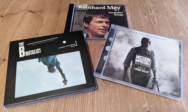 Three CDs lying on a wood desk plate. 
In the foreground: A black CD case with an upside-down picture of the upper body of the Statue of Liberty and "The Brutalist" – Original Motion Picture Soundtrack – By Daniel Blumberg" written on it; and a white/greyish CD cover showing the grey and black silhouette of a man, "Suites and Themes from the Motion Picture" written at the top and " "Tom Cruise – Mission: Impossible – Dead Reckoning Part One – Music by Lorne Balfe".
In the background: A CD cover showing a slightly tanned man with short hair, "Reinhard Mey – Die großen Erfolge" ("Reinhard Mey – The big successes") written above/next to the man's hair.