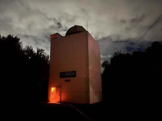 The Westport Observatory stands illuminated with a red light glowing from its entrance under a cloudy sky.