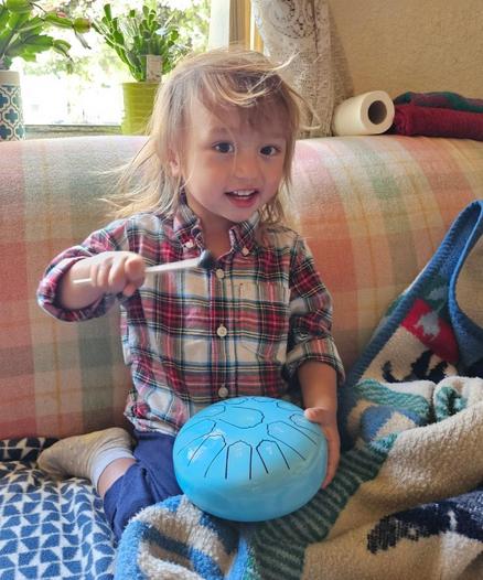 A young child, wearing a red and blue plaid shirt and blue pants, seated on a couch with a multicolored plaid blanket, is holding a bright blue, round handpan drum with etched lines and numbers, a drumstick poised to strike it; the child smiles at the camera, and light enters from a window in the background.