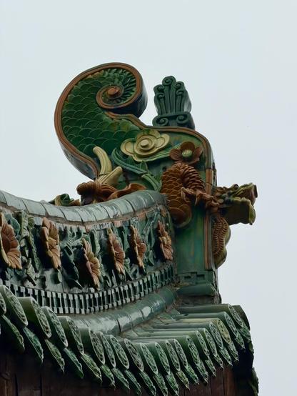 Close-up of an ornate roof corner from traditional Chinese architecture, featuring green glazed tiles and intricate ceramic decorations shaped like flowers, scales, and a dragon head against a pale sky.