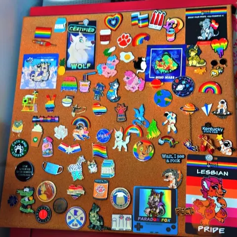 A cork board with enamel pins scattered randomly on it. Some include rainbow lgbtq pride ones, pins from Fursona Pins, astronaut pins, and My Little Pony ones.