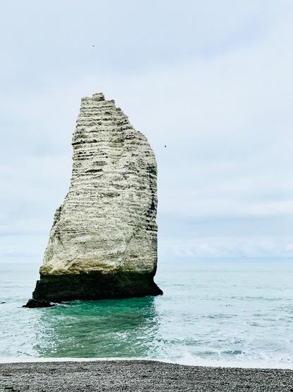 Ein einzelner heller Felsen ragt steil aus dem türkisblauen Meer empor. Seine strukturierte Oberfläche zeigt Schichten aus Kalk und Stein, während die Wellen ruhig an seiner Basis brechen. Der Himmel ist leicht bedeckt und verleiht der Szene eine klare, maritime Ruhe.