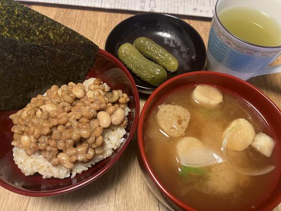 A bowl of rice topped with natto (fermented soybeans) and a sheet of nori. Beside it, a bowl of miso soup with translucent pieces, and a plate of pickled cucumbers, along with a cup of green tea